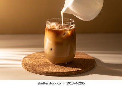 Milk cream is poured into a iced cold brew coffee. Coffee cold cocktail drink with ice and milk in morning sun light.