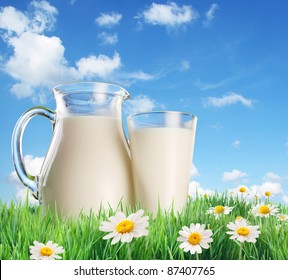 Milk jug and glass on the grass with chamomiles. On a background of the summer sky with clouds.