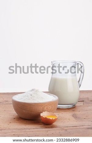 milk in jug with wooden bowl with white flour and cracked egg on rustic wooden table and white background.