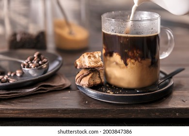 Milk Pouring A Swirl Into A Clear Glass Cup Of Coffee. Two Biscotti Sit Atop A Black Saucer With The Mug. A Scoop Of Coffee Beans Are On The Side, Brown Sugar And Coffee Are Blurred In The Background.