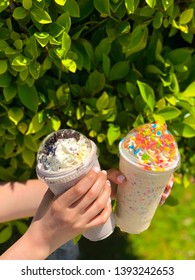 Milk Shakes On A Sunny Day. Fruity Pebble On Milkshake With A Green Background On A Summer Day. A Young Girl With Nice Nails Holding Milkshakes. Delicious Food.