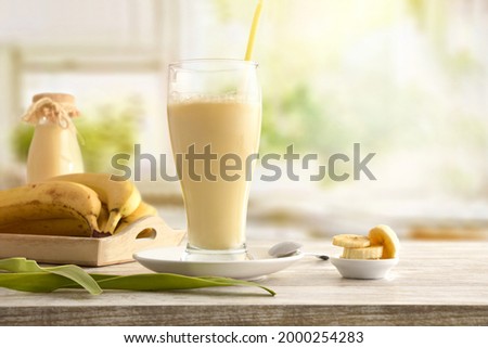 Milkshake with banana in glass cup on table with tray with fruit and kitchen background. Front view.