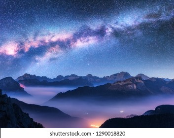 Milky Way above mountains in fog at night in autumn. Landscape with alpine mountain valley, low clouds, purple starry sky with milky way, city illumination. Aerial. Passo Giau, Dolomites, Italy. Space