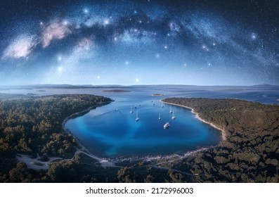 Milky Way Arch And Beautiful Yachts And Boats On The Sea Bay At Summer Night. Kamenjak, Croatia. Space. Top View Of Arched Milky Way, Starry Sky, Sailboats, Lagoon, Clear Blue Water, Forest. Travel