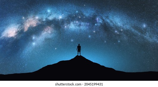 Milky Way Bogen und Mann auf dem Berggipfel in der Sternennacht. Silhouette des Alleinstehenden, blauer Himmel mit hellen Sternen im Sommer. Galaxie. Weltraumhintergrund. Landschaft mit gewölbter Milchstraße. Reisen und Natur