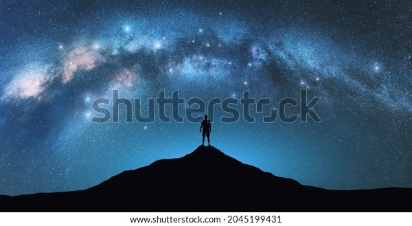 Milky Way arch and man on the mountain peak at
starry night. Silhouette of alone guy, blue sky with bright stars
in summer. Galaxy. Space background. Landscape with arched milky
way. Travel and nature