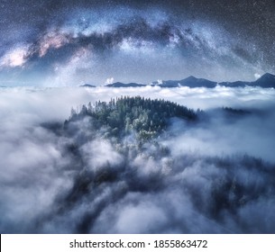 Milky Way Arch Over The Mountains In Low Clouds At Starry Night In Summer. Landscape With Sky With Stars, Arched Milky Way, Trees On The Hill In Fog, Mountain Peaks. Space And Galaxy. Aerial View  