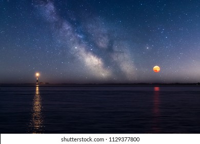 The Milky Way and Full moon in the sky with a Lighthouse beacon reflecting in the water. 