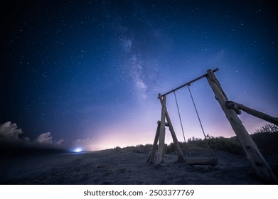 The Milky Way stretches across the night sky. A solitary wooden swing set stands on a sandy beach, inviting viewers to imagine swinging under the vast, starry sky. Zhuangwei, Yilan County, Taiwan. - Powered by Shutterstock