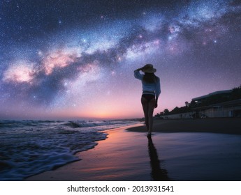 Milky Way and young woman in hat on sandy beach against purple starry sky reflected in water at night. Landscape. Summer travel. Girl on the tropical sea coast and bright Milky Way and stars. Space