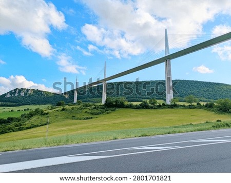 The Millau Viaduct, a large cable-stayed bridge, spans across a green valley with hills and fields in Millau, France