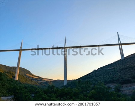The Millau Viaduct, a large cable-stayed bridge, spans across a green valley with hills and fields in Millau, France