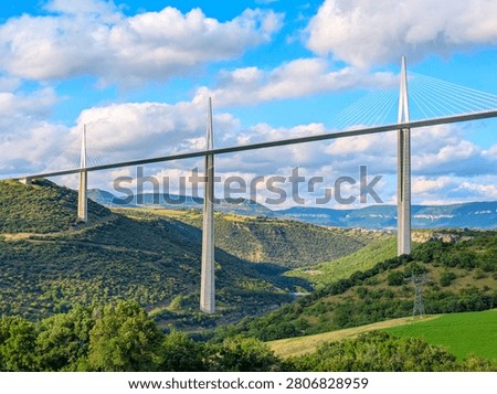 The Millau Viaduct, a large cable-stayed bridge, spans across a green valley with hills and fields in Millau, France