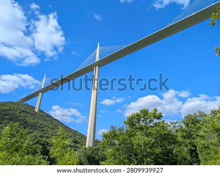 The Millau Viaduct, a large cable-stayed bridge, spans across a green valley with hills and fields in Millau, France