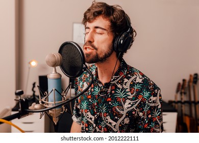 Millenial Male Singer On Headphones Singing Into Microphone While Working On Songs During A Recording Studio Session At Home.