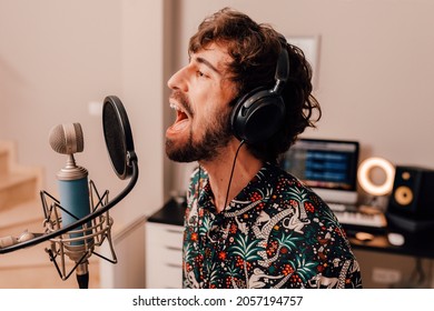 Millenial Male Singer On Headphones Singing Into Microphone While Working On Songs During A Recording Studio Session At Home.