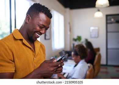 A Millenial Man Smiles And Looks At His Phone In The Office With Colleagues