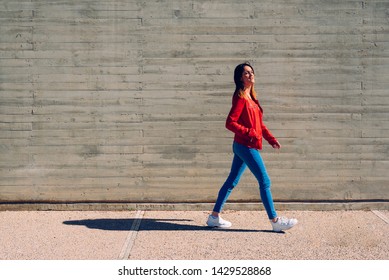 Millenial woman walking calmly in blue and red jeans next to a cement wall, gray background copy space.
