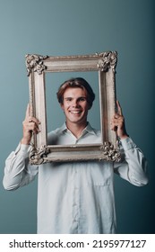 Millenial Young Man Artist With Blonde Hair On Gilded Picture Frame Portrait. Isolated On Blue Background In Studio.