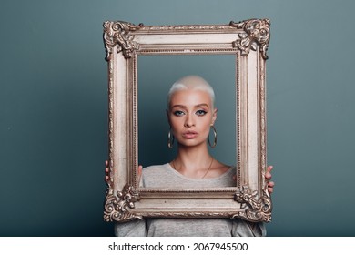 Millenial Young Woman With Short Blonde Hair Holds Gilded Picture Frame In Hands Behind Her Face Portrait