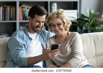 Millennial adult son demonstrates to older mother new application on smartphone sit on couch at home, helps her and teaches, shows old mom photos, share personal, having fun, modern tech usage concept