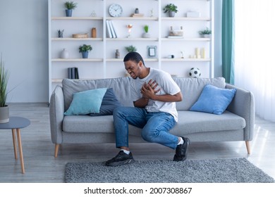 Millennial African American Guy Having Heart Attack, Touching His Chest In Pain, Sitting On Sofa At Home, Full Length. Young Black Man Suffering From Cardiovascular Ache, Getting Infarction Indoors