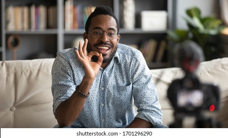 Millennial African American Male Blogger Or Tutor Sit On Couch Shooting Video Blog On Modern Web Cam, Young Biracial Man Coach In Glasses Talk With Subscribers Record Tutorial Or Training Course