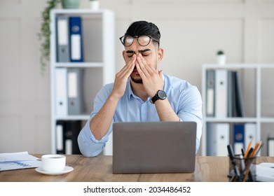 Millennial Arab Employee Rubbing Tired Irritated Eyes, Working With Laptop Too Much, Exhausted From Online Job At Office. Young Eastern Businessman Feeling Fatigue From Looking At Screen