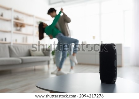 Millennial Asian couple dancing to popular music at home, selective focus on portable wireless speaker on table, copy space. Young family moving to their favorite song, using modern device