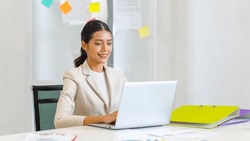 Millennial Asian Happy Cheerful Pretty Businesswoman Secretary Employee Sitting Smiling Enjoy Working Online Typing Laptop Computer At Workstation Desk Full Of Document Folders In Company Office.