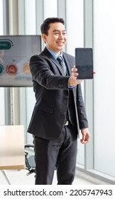 Millennial Asian Happy Successful Professional Male Businessman Entrepreneur In Formal Suit Standing Holding Touchscreen Tablet Computer Smiling In Company Office Meeting Room With Big Screen Monitor.