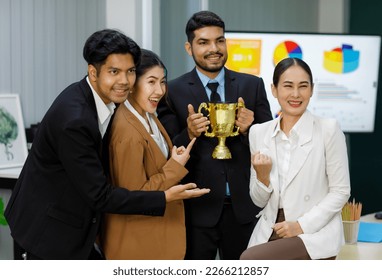 Millennial Asian Indian professional successful bearded businessman in formal suit smiling winning holding business competition golden reward trophy while male female colleagues celebrating together. - Powered by Shutterstock