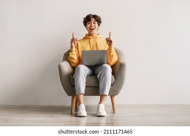 Millennial Asian man having creative idea, gesturing eureka with both hands, pointing up, offering space for design, sitting in armchair with laptop pc against white studio wall, copy space - Shutterstock ID 2111171465