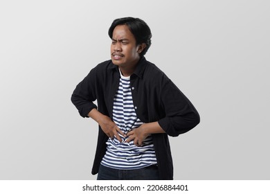 Millennial Asian Man Suffering From Stomach Ache, Holding Belly, Feeling Discomfort. Upset Man With Abdomen Pain, Gastritis Symptom, Indigestion Problem, Isolated On White Studio Background