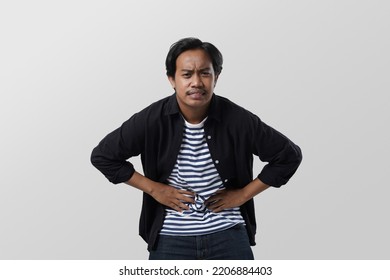 Millennial Asian Man Suffering From Stomach Ache, Holding Belly, Feeling Discomfort. Upset Man With Abdomen Pain, Gastritis Symptom, Indigestion Problem, Isolated On White Studio Background