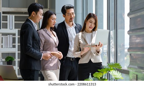 Millennial Asian Professional Successful Businesswoman In Formal Suit Standing Holding Showing Laptop Notebook Computer Screen Discussing With Male And Female Colleagues In Company Office Workstation.