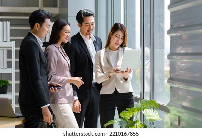 Millennial Asian Professional Successful Businesswoman In Formal Suit Standing Holding Showing Laptop Notebook Computer Screen Discussing With Male And Female Colleagues In Company Office Workstation.