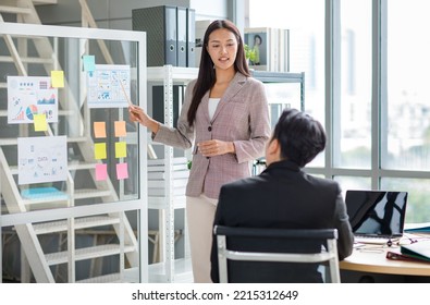 Millennial Asian professional successful female businesswoman presenter lecturer speaker in formal suit standing using pencil pointing graph chart document presentation on glass board in meeting room. - Powered by Shutterstock