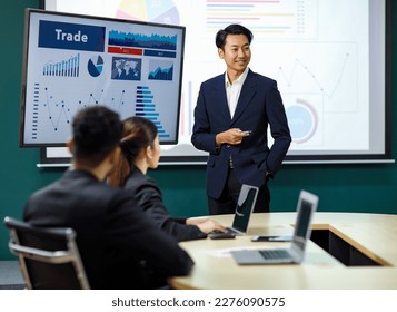 Millennial Asian professional successful male businessman presenter speaker lecturer in formal business suit hold pen point trading buy sell presentation monitor present to female audiences in office. - Powered by Shutterstock