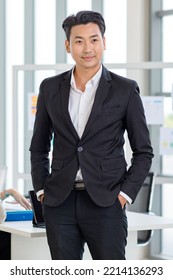 Millennial Asian professional successful young handsome businessman in formal suit standing posing put hands in pants pockets while male female colleagues discussing in company office meeting room. - Shutterstock ID 2214136293