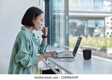 Millennial Asian student prepares for test or exam browses laptop studies in coffee house makes notes in notepad watches online webinar learns foreign language. E learning and online studying