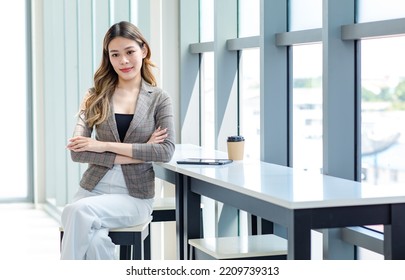 Millennial Asian Successful Professional Female Businesswoman In Formal Suit Take Break Sitting Posing Crossed Arms Uesing Tablet Computer And Drinking Coffee From Disposable Cup In Office Room