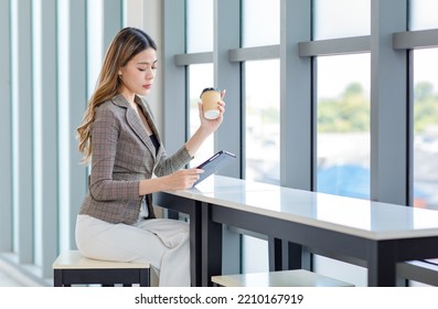 Millennial Asian successful professional female businesswoman in formal suit take break sitting smiling browsing surfing internet via tablet computer and drinking coffee from disposable cup in office. - Powered by Shutterstock