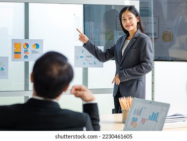 Millennial Asian successful professional female businesswoman lecturer in formal suit standing smiling pointing finger at graph chart data information on glass board presenting explaining to audience. - Powered by Shutterstock