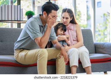 Millennial Asian Unhappy Family Mother Sitting On Sofa Couch Holding Hugging Soothing Little Young Female Daughter While Sad Stressed Father Crying After Big Fight Argument In Living Room At Home.