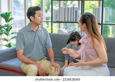 Millennial Asian Unhappy Family Mother Sitting On Sofa Couch Holding Hugging Soothing Little Young Female Daughter While Sad Stressed Father Crying After Big Fight Argument In Living Room At Home.