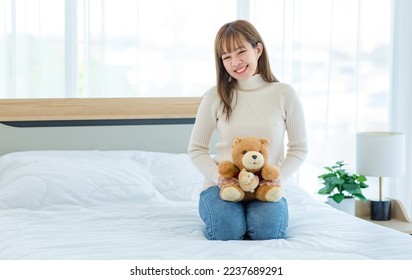 Millennial Asian young beautiful female teenager model in casual outfit turtleneck sweater and jeans sitting on bed smiling holding two little cute teddy bear dolls present gift in bedroom in morning.