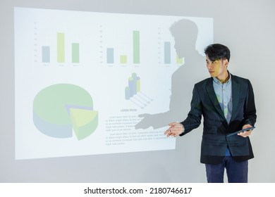 Millennial Asian young cheerful male businessman presenter in formal suit standing holding notebook and pen pointing presenting corporate graph chart data information from wall screen in meeting room. - Powered by Shutterstock