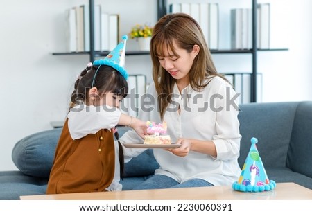 Millennial Asian young female teenager mother nanny babysitter sitting holding dish while little cute preschooler daughter birthday girl wearing tall hat feeding cake with hand to mom mouth in party.