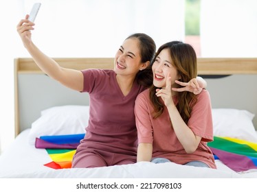 Millennial Asian Young Happy Cheerful Female Teenager LGBTQ Lesbian Couple Sitting Smiling Together On Bed Holding Two Fingers Peace Sign Under Blanket And Rainbow Flag Taking Selfie With Smartphone.